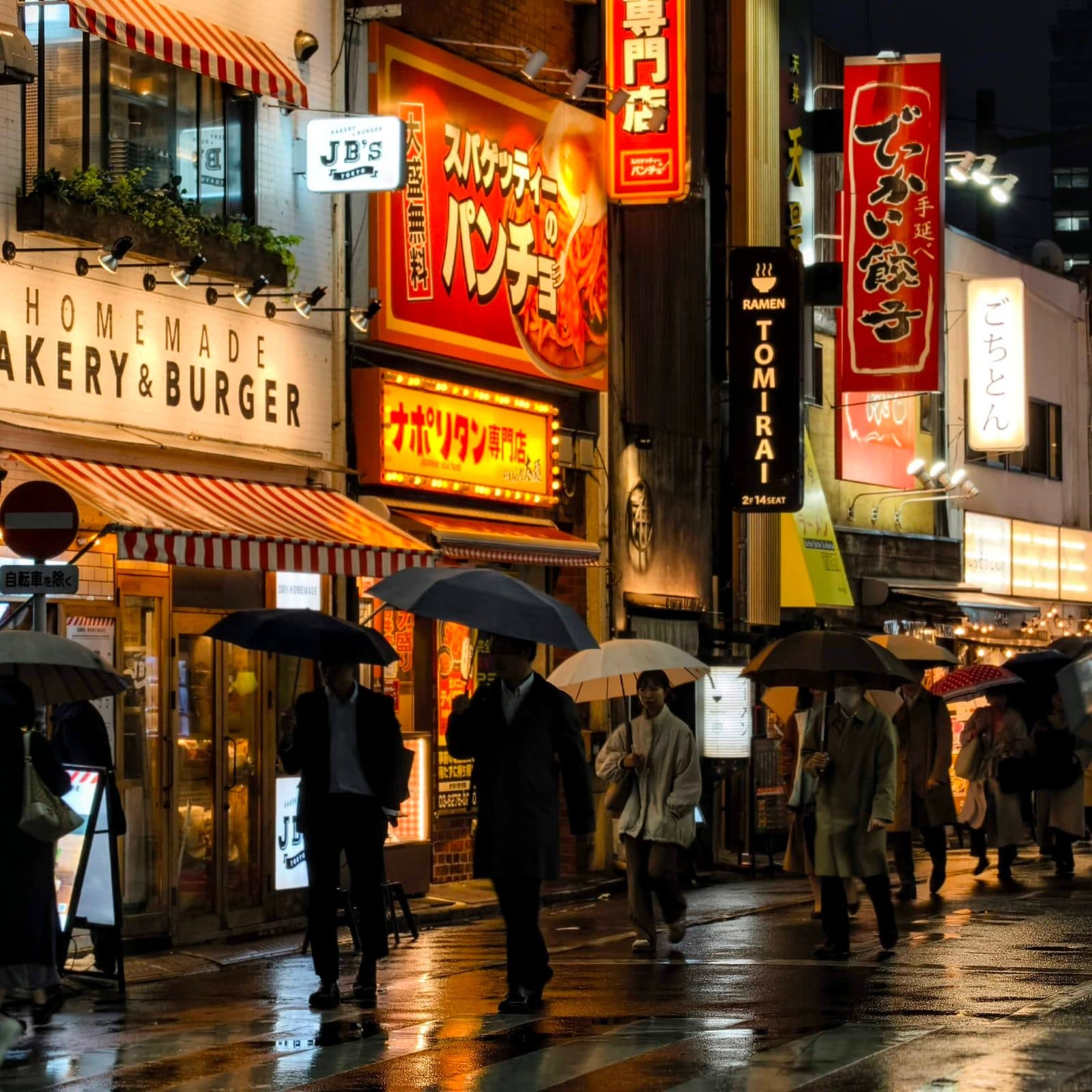 A Tokyo street at night in the rain, with people under umbrellas and lit signs overhead