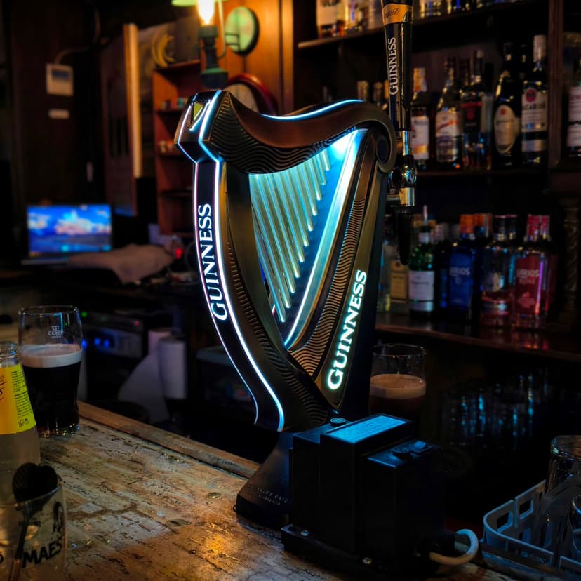 A lit-up Guinness harp tap on a worn wooden bar counter, with a half-poured pint settling beside it and shelves of bottles behind